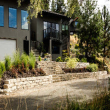 Load image into Gallery viewer, A large, scale retaining wall made of gray concrete stones, installed in a garden setting with greenery and a house in the background.