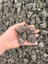 Load image into Gallery viewer, Hand holding a small pile of dark rocks against a background of similar rocks
